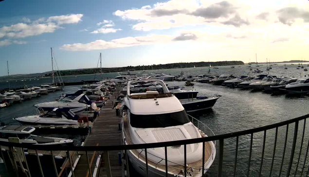 A marina filled with numerous boats and yachts docked along wooden piers, with calm water and a partly cloudy sky overhead. In the distance, there is a shoreline with additional boats and some landforms.