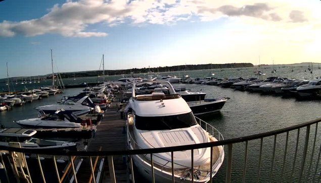 A marina with numerous boats and yachts docked along wooden piers, under a partly cloudy sky during sunset or sunrise, with calm water extending to a distant shoreline.