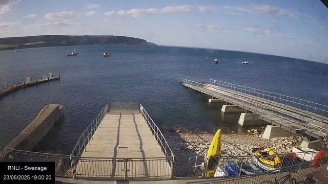 A view of a harbor with a concrete pier extending into the water. There are several boats floating on the calm sea. In the foreground, there is a yellow life raft and some equipment on a rocky shoreline. The sky is partly cloudy with patches of blue. The background features a headland with cliffs. The timestamp indicates it was taken on June 23, 2025, at 19:00.
