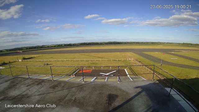 A view from a webcam at Leicesterhire Aero Club showing an empty airport apron with a windsock and a small fenced area. In the background, there is a runway with some markings, grassy areas, and a partly cloudy sky with the time and date displayed in the top right corner: 23-06-2025 19:00:21.
