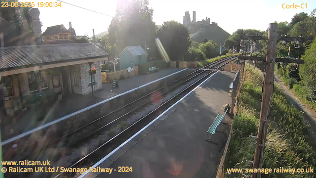 A small railway station platform bathed in sunlight. There is a building with a tiled roof on the left side, next to a flower basket. An "WAY OUT" sign is visible near the building. Beside the platform, there are train tracks stretching into the distance, curving slightly to the right. A row of wooden fencing lines the far side of the platform, and two benches are placed on the right side. Overhead, electrical wires and poles run parallel to the tracks. In the background, lush green trees and a hilltop castle ruins are visible, with the sun shining brightly, creating lens flare effects. The scene appears calm and quiet.