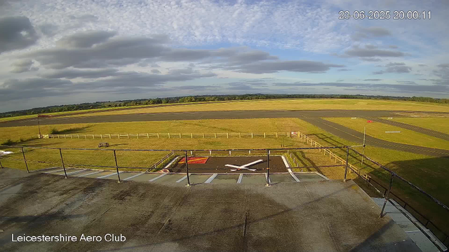 A view from a webcam at Leicestershire Aero Club showing an empty helicopter landing pad with a fenced perimeter. Beyond the pad, there are grassy fields, taxiways, and runways stretching into the distance. The sky is partly cloudy with patches of blue, and the timestamp indicates it was taken on June 23, 2025, at 8:00 PM.