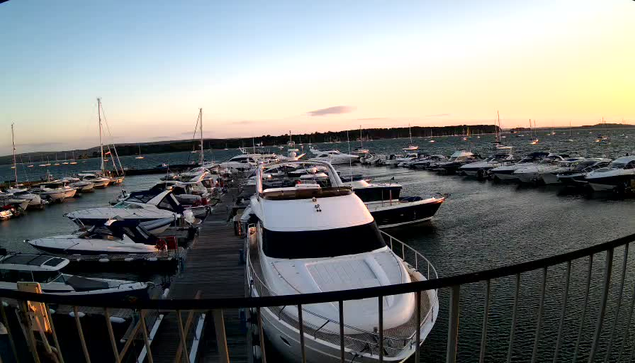 A marina filled with numerous boats and yachts docked along wooden piers during sunset, with calm water reflecting the sky's soft light and a distant shoreline in the background.