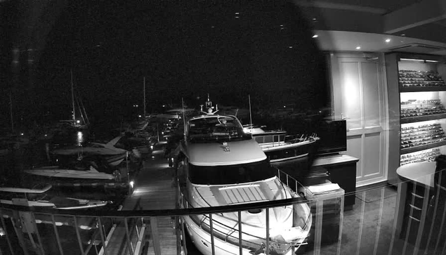 A black-and-white photo taken through a window showing a marina at night, with multiple yachts and boats docked closely together. The scene includes a railing in the foreground, and a well-lit interior space with shelves or display cases on the right side.