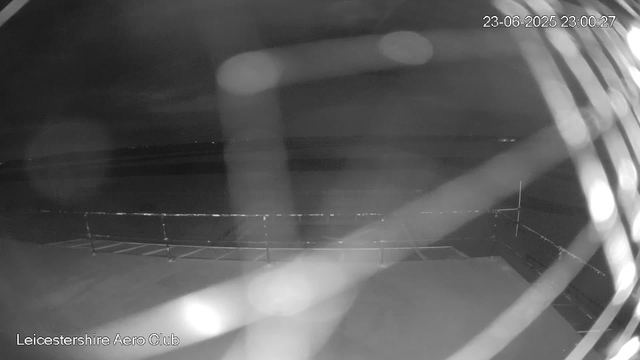 A black-and-white image from a webcam shows an outdoor scene at night. The area appears to be an open space with a fence or railing in the foreground. The background is dark, with no visible lights or detail. The timestamp in the top right corner reads "23-06-2025 23:00:27," and text in the bottom left corner indicates the location as "Leicestershire Aero Club."