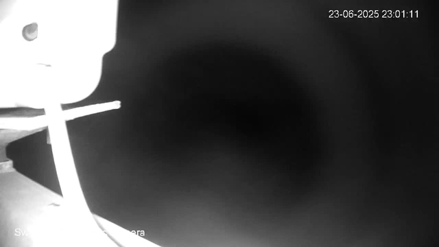 A black and white webcam image shows part of a table or surface on the left side, with a blurred object on top. The majority of the image is dark, with a faint circular shape in the center. In the upper right corner, the date and time are displayed: 23-06-2025 23:01:11.