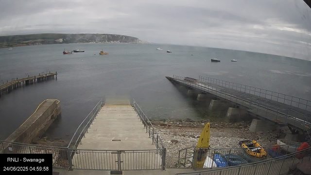 A view of a harbor with a cloudy sky. There are boats floating on the water and a pier extending into the harbor. In the foreground, there are two concrete docks, a yellow caution sign, and a small yellow boat inside a fenced area. The background features a rocky coastline with cliffs and some buildings. The date and time displayed are 24/06/2025 at 04:59:58.