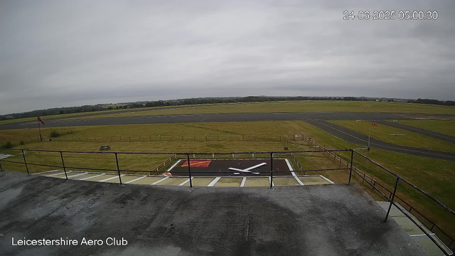 A view from a webcam at Leicestershire Aero Club showing an empty aircraft parking area in the foreground with a large, empty helipad featuring a white "X" marker, and a runway in the background stretching across a mostly cloudy sky during daytime. The landscape is flat with open grassy areas and some distant trees. The date and time on the image are 24th June 2025 at 5:00 AM.