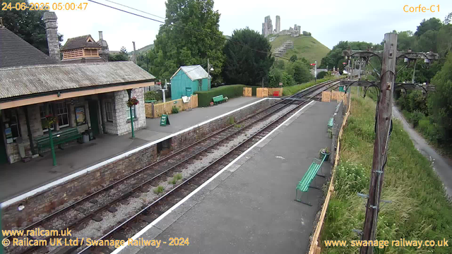 A small railway station platform with a stone building that has a sloped roof and a chimney on the left. There are green benches, potted flowers, and hanging baskets on the platform. A green bin is next to the building, and there's a small turquoise shed in the background. The railway tracks run through the center of the image, with a safety barrier at the far end. On the right side, there are wooden utility poles with wires, a narrow footpath with grassy vegetation beside it, and benches along the platform. In the background, a hill with ruins or a castle is visible, surrounded by lush green trees, and the sky is overcast. The timestamp in the top left corner shows "24-06-2025 05:00:47," and there are website credits in the bottom corners.