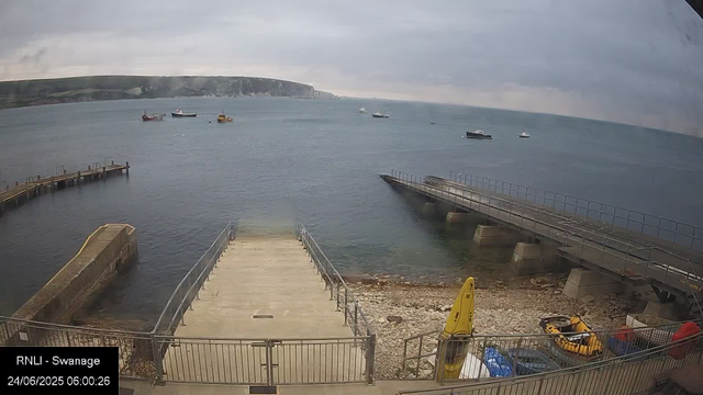 A coastal scene showing a small pier extending into calm waters, with a separate floating walkway to the right. In the foreground, there are some yellow and orange safety equipment, a fenced area, and a rocky beach. Several boats are anchored in the water further out, and a distant headland with cliffs is visible under an overcast sky.