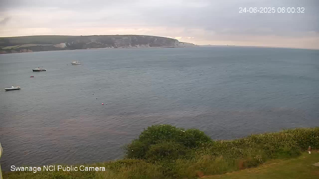 A view of a calm sea with several small boats floating on the water, set against a backdrop of white chalk cliffs and green hills under a cloudy sky. Some greenery is visible at the bottom of the image. The timestamp in the upper right corner indicates the date and time as 24-06-2025 06:00:32.