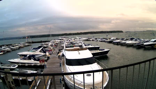 A marina with numerous docked boats and yachts extending into calm water. The photo is taken from a higher vantage point, with a black railing visible in the foreground. The sky is partly cloudy with a soft, pastel hue, suggesting either dawn or dusk.