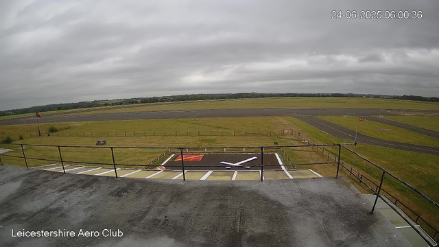 The image shows an empty helicopter landing pad with white boundary lines and a large red H symbol in the center. There is a railing around the edge of the platform. In the background, there is a grassy area with a windsock on a pole and a small bench. Beyond that, there are grass fields and a runway with markings, under a cloudy sky. The timestamp in the top right corner reads "24-06-2025 06:00:36," and the bottom left corner has the text "Leicestershire Aero Club."