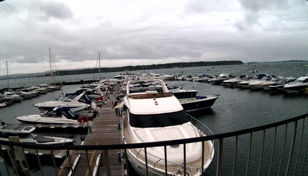 A marina filled with numerous boats and yachts docked along wooden piers under a cloudy sky, with calm water and a distant shoreline in the background.