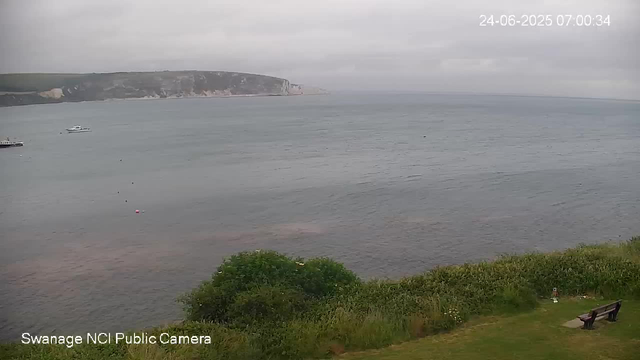 A view of a calm sea with a distant headland in the background. The sky is cloudy, and a few boats are floating on the water near the shoreline. In the foreground, there is a grassy area with some bushes and a bench facing the water. The date and time stamp in the top right corner reads "24-06-2025 07:00:34."
