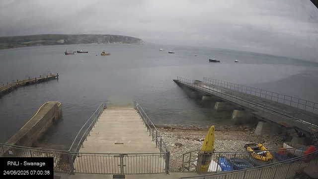 A view of a harbor with a dock in the foreground, leading out to calm water. Several boats are anchored across the bay, and a distant cliff rises along the shoreline under a cloudy sky. There are some equipment and safety devices near the dock area. The timestamp indicates the photo was taken on June 24, 2025, at 7:00 AM.