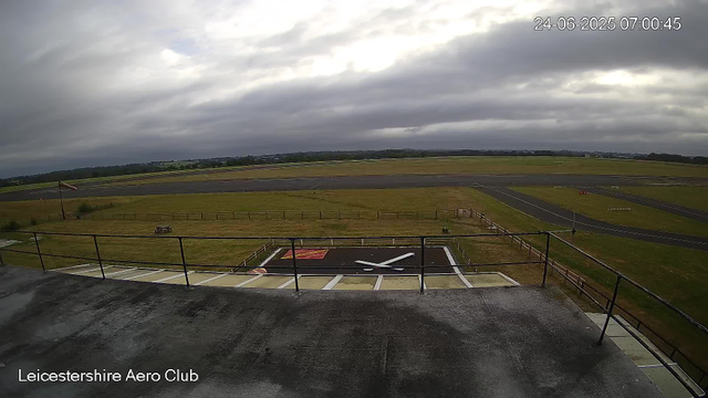 A view from a webcam at Leicestershire Aero Club shows a cloudy sky over an airfield with runways stretching into the distance. In the foreground, there is a rooftop area with a small helipad marked with an "H" and a windsock. The scene appears calm with no aircraft present. The timestamp indicates it is early morning on June 24, 2025.