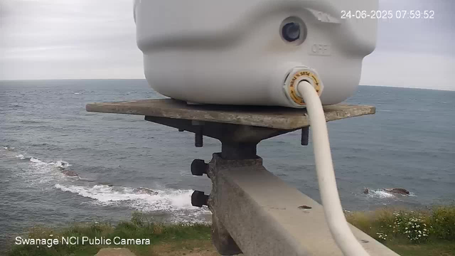 A close-up view of a weather station or equipment mounted on a wooden platform, overlooking the ocean. The ocean waves are visible in the background beneath an overcast sky. The image includes a timestamp in the upper right corner showing "24-06-2025 07:59:52," and a label in the lower left corner reading "Swanage NCI Public Camera."