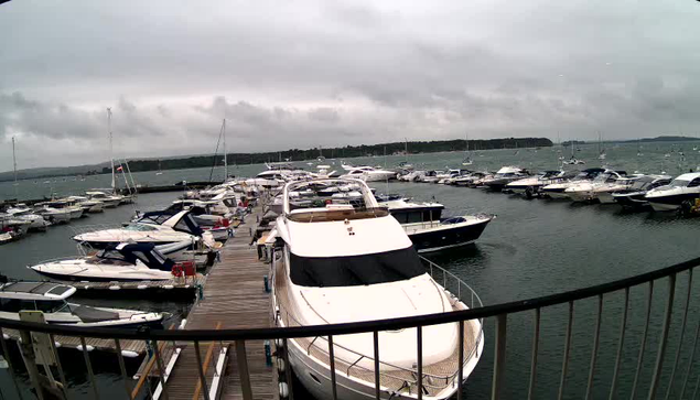 A marina with numerous boats docked along a wooden pier under a cloudy, overcast sky. The boats vary in size and style, and some have covers or sails. The water appears calm, and distant land with more boats can be seen on the horizon.