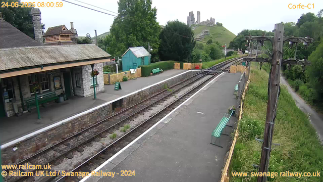 The image shows a small, quiet railway station platform with two train tracks running through it. There is a stone building with a sloped roof and green benches on the platform. A small teal-colored shed and a green hedge are behind the benches. To the right, there are yellow safety barriers along the edge of the platform. A wooden utility pole with multiple wires stands beside the tracks. In the background, across the tracks, there is a grassy hill with a medieval castle ruin visible at the top. The surrounding area is lush with trees and greenery, and a small path runs parallel to the railway tracks on the right side. The timestamp in the top left corner reads "24-06-2025 08:00:59," and the image credits indicate it is from a webcam at Swanage Railway.