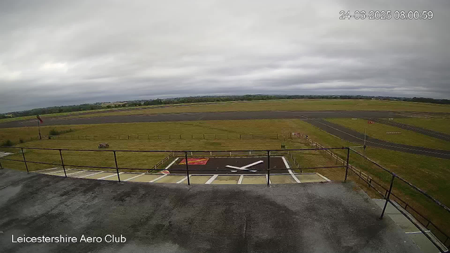 A view from a webcam at Leicestershire Aero Club shows an overcast sky with gray clouds. In the foreground, there is a small helipad with a white "H" symbol and red markings, surrounded by a metal railing. The grass area around the helipad is open, with a few small objects visible. Beyond the grassy area, there is a runway with dark asphalt, extending horizontally across the image. The landscape is flat, with fields and trees visible in the distance under a cloudy sky. The timestamp shows the date as June 24, 2025, at 8:00 AM.