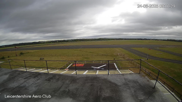 A view from a webcam at Leicesterhire Aero Club showing an overcast sky with dark clouds. In the foreground, there is a black helipad with a white helicopter landing pad marking. The ground has a wet appearance, indicating recent rain. Beyond the helipad, there is a grassy area with a small flagpole on the left side. In the background, there are empty paved runways and taxiways stretching across the landscape, with a distant tree line and fields under the cloudy sky. The timestamp indicates the image was captured on June 24, 2025, at 09:00:29.