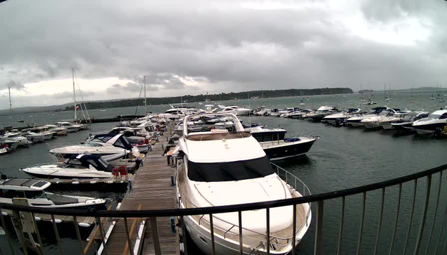 A marina filled with various boats and yachts docked along a wooden pier. The sky is cloudy and overcast, with dark clouds hanging above the water. In the distance, there is a tree-lined shoreline.