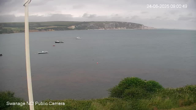 A view of a calm body of water with four boats floating on the surface, some distance from the shore. In the background, there are white cliffs and green hills under a cloudy sky. In the foreground, there is green shrubbery and part of a white pole on the left side. The image has a timestamp in the upper right corner showing "24-06-2025 09:00:49", and a label in the lower left corner indicating it is from "Swanage NC Public Camera."