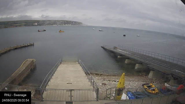 A view of a small harbor with a concrete pier extending into calm water. To the right, there is a metal ramp leading into the water, supported by concrete piers. On the shore, there is a yellow caution sign and some equipment including a yellow inflatable boat and blue containers. Several small boats are anchored in the water, and a hilly coastline with cliffs is visible in the background under a cloudy sky.
