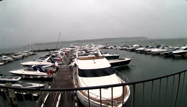 A marina filled with numerous white and dark-colored yachts and boats docked along wooden piers, with a cloudy sky overhead and a distant shoreline visible in the background.