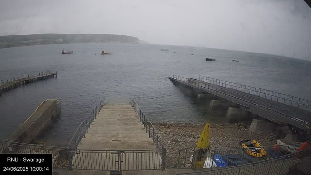 A view of a cloudy day at a small harbor with a concrete pier extending into the water. Several boats are anchored in the distance, and a larger landmass can be seen on the horizon. The shoreline has some equipment and a yellow life buoy, with a metal railing along the pier. The date and time displayed are June 24, 2025, at 10:00:14.