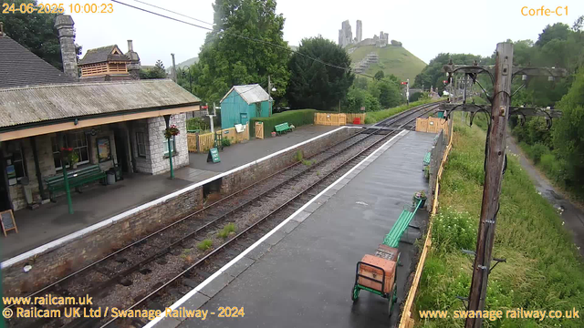 A small railway station platform with a stone building on the left, featuring hanging flower baskets and benches. The railway tracks run through the center of the image, with some weeds sprouting between the rails. A green bench and a small blue shed are visible on the far side of the platform. There are wooden fences and a few decorative plant containers along the platform's edge. To the right, there is a tall wooden utility pole with electrical wires extending from it. A narrow gravel footpath runs alongside the platform, bordered by lush green bushes and trees. In the background, a hill with the ruins of a castle or fortress is visible, partially obscured by mist or fog. The image has the date and time stamped as June 24, 2025, at 10:00:23.