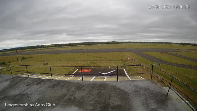 A view from a webcam at Leicestershire Aero Club showing an overcast sky with grey clouds, a runway in the distance, and a small helipad or landing area with a painted white letter "X" on the ground in the foreground. The landscape is mostly grassy fields with some fencing, and there are no people or aircraft visible in the image. The timestamp indicates it was taken on June 24, 2025, at 10:00 AM.