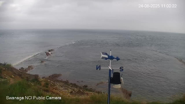 An image shows a view of the ocean from a coastal area on a cloudy day. In the foreground, there is a weather vane with an arrow and directional markers indicating north and south. The water appears calm with gentle waves, and the coastline includes some rocks and low vegetation. The timestamp in the top right corner reads "24-06-2025 11:00:22."