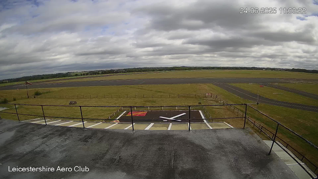 A view from a webcam at Leicesterhire Aero Club shows an overcast sky with thick gray clouds. In the foreground, there is a section of a building with a raised platform and a railing. On the ground, there is a helicopter landing pad marked with a large "H" and a red circle around it. The airfield extends into a grassy area with paved taxiways and runways. In the distance, there are open fields and some scattered trees under the cloudy sky. The timestamp in the top right corner indicates the date as June 24, 2025, and the time as 11:00:22.