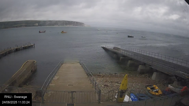 A view of an overcast day at the harbor in Swansea, showing the sea with several boats floating, a wooden dock extending into the water, and a sloped walkway leading down to the rocky shoreline. In the foreground, there are some yellow safety signs and equipment, with a fenced area surrounding the scene. The date and time are displayed as June 24, 2025, at 11:00:24.