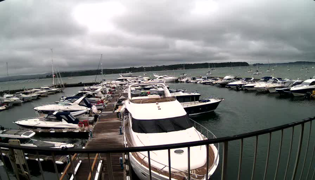 A marina filled with numerous boats and yachts docked along the wooden piers, under a cloudy sky. The water is calm, and the boats vary in size and style, with some having covered cabins. A railing is visible in the foreground, indicating the photo was taken from a vantage point overlooking the marina. In the background, there is a distant shoreline with land and trees.