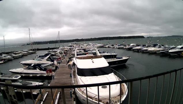 A marina filled with various boats and yachts docked along a wooden pier. The sky is overcast with gray clouds, and distant land is visible across the water. The boats are mostly white, and the scene appears calm with gentle waves.