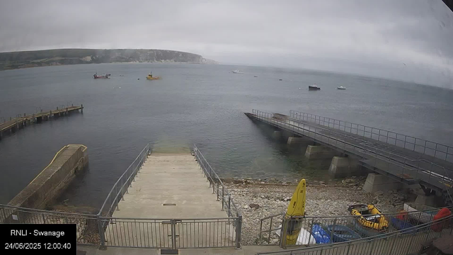 A view of a small harbor on a cloudy day with calm water. There are boats floating in the distance, a pier extending into the water, and a ramp leading into the sea. In the foreground, there are some yellow and red safety equipment and a yellow caution sign on the rocky shore. A fence surrounds the area, and the timestamp indicates it is midday on June 24, 2025.