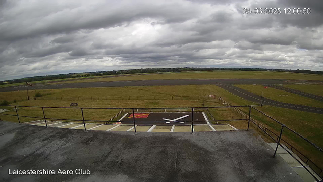 A view from a webcam at Leicesterhire Aero Club showing a cloudy sky over a runway and grassy fields. The time and date are displayed as 24-06-2025 at 12:00:50. The foreground includes a small helipad area with a white helicopter silhouette painted on the ground, and a railing surrounds the viewing platform. The horizon shows open fields and trees under dark, overcast clouds.