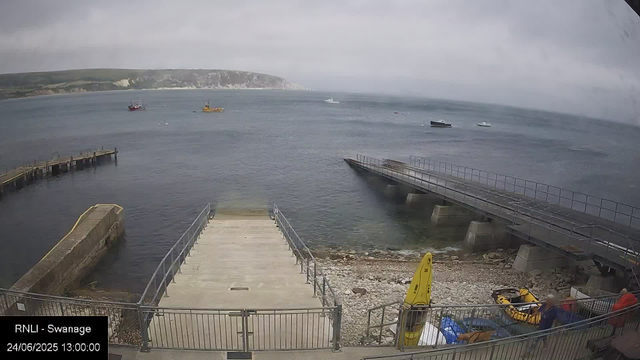 A view of a harbor with a concrete pier extending into the water, surrounded by a metal railing. To the right, there is a ramp or slipway with a metal railing, leading down into the water. Near the bottom right corner, there is a yellow rescue buoy and some orange equipment. Two people are standing near the railing, one in a red jacket and another in a blue jacket. The water is calm with several boats floating in the distance, and a hilly coastline with greenery and white cliffs can be seen under a cloudy sky.