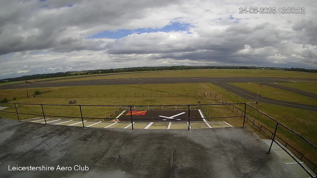 A view from a webcam showing an airfield with a paved runway and surrounding grassy areas. The sky is partly cloudy with some patches of blue. In the foreground, there's a helipad marked with a white "H" on a black surface, with a red square nearby. The image timestamp reads "24-06-2025 13:00:02," and the location label in the bottom left corner says "Leicestershire Aero Club."