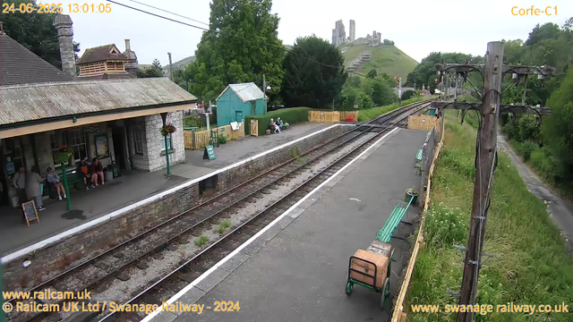 A small train station platform with a few passengers sitting and waiting. There are green benches, a small teal-colored shed, and a yellow picket fence surrounding the area. Above, there are power lines and an old wooden telegraph pole. In the background, a hill with a historic castle or ruins atop it is visible, along with lush green trees and hills. The sky is overcast, and the scene appears calm and quiet. The timestamp indicates it was taken on June 24, 2025, at 1:01 pm.