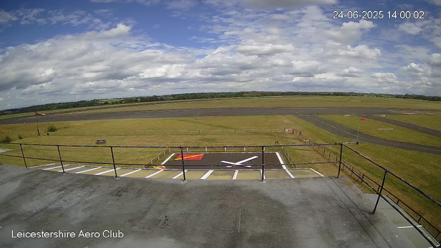 This image shows a view from a webcam at Leicesterhire Aero Club during the daytime. In the foreground, there is a landing pad with markings and a small red and yellow painted area. A windsock on a pole is visible to the left, indicating wind direction. The background features a runway with open fields on either side, and a partly cloudy sky overhead. The timestamp in the top right corner reads "24-06-2025 14:00:02."