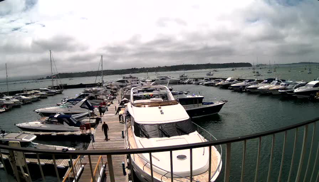 A view from a balcony overlooking a marina filled with various boats and yachts docked along piers. The sky is overcast with gray clouds, and the water appears calm. The scene includes a large white yacht in the foreground and several smaller boats on either side. In the background, there is a distant shoreline with some boats on the water.