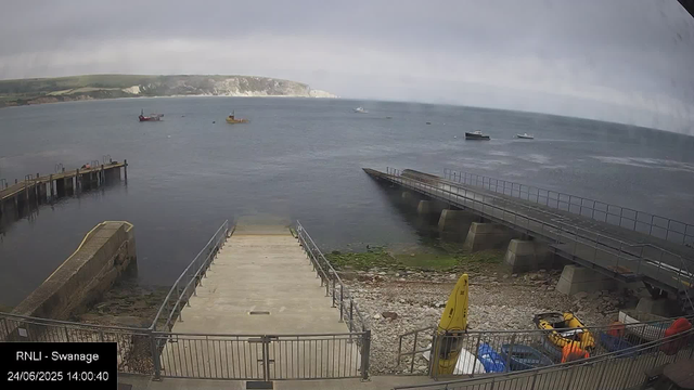 A view of a harbor with a concrete pier in the foreground, surrounded by calm water. Several boats are anchored in the distance, and a grassy cliff with white chalk cliffs is visible on the far side of the bay. The sky is overcast with grey clouds. The timestamp indicates the date and time as 24/06/2025 at 14:00:40.