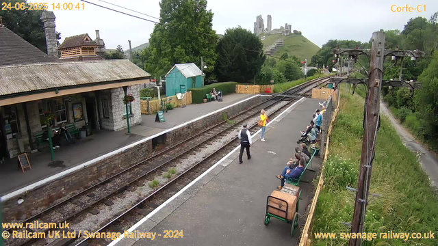 A small train station platform with several people waiting. Some are sitting on green benches, while others are standing. A person in a yellow jacket is walking along the platform, and a train conductor or station staff member is walking toward them. There are a few bicycles parked near the benches. The station building is made of stone with a sloped roof, and hanging flower baskets decorate the entrance. A small green shed is located behind the platform. In the background, there is a grassy hill with the ruins of a castle on top, and a railway line with overhead electrical wires runs along the platform. A utility pole with wires is visible on the right side. The overall scene is lush with green trees and vegetation surrounding the station. The timestamp indicates the image was captured on June 24, 2025, at 14:00.