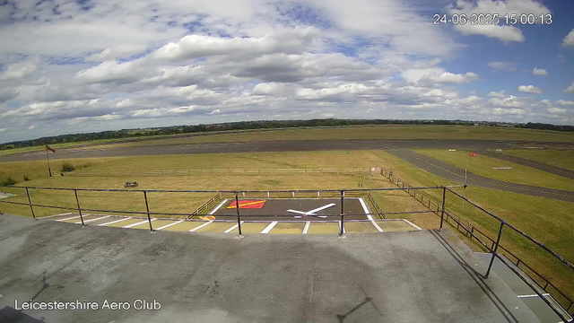 A view from a webcam at Leicestershire Aero Club shows a partly cloudy sky over an airfield with grassy areas, a small windsock, and a tarmac runway in the distance. In the foreground, there is a rooftop with a railing, painted markings, and a large taxiway or helipad symbol. The time stamp indicates it was taken on June 24, 2025, at 15:00.
