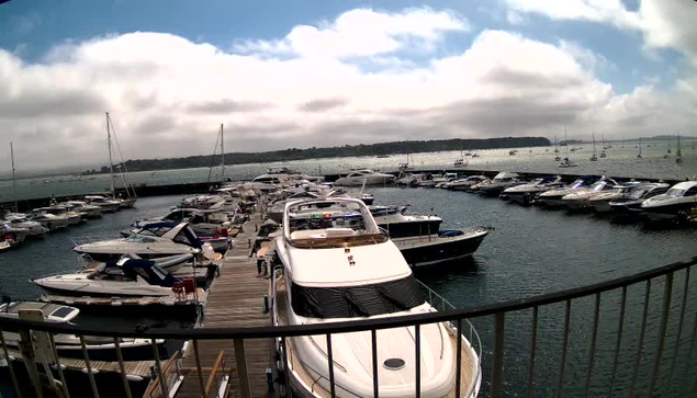 A view of a marina filled with various boats and yachts docked along wooden piers under a cloudy sky. The photo is taken from a balcony or viewing platform with a black railing in the foreground.
