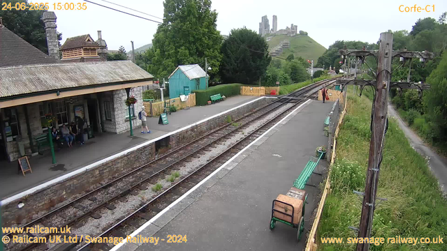 A small railway station platform with a few people waiting. There are green benches, a small teal-colored building, and a yellow fence. A large utility pole with electrical wires is on the right side of the image. In the background, a castle is perched on a hill surrounded by greenery. The time and date are displayed in the upper left corner: June 24, 2025, at 15:00:35. The surrounding area includes trees, a pathway on the right with overgrown grass, and a small building on the left.